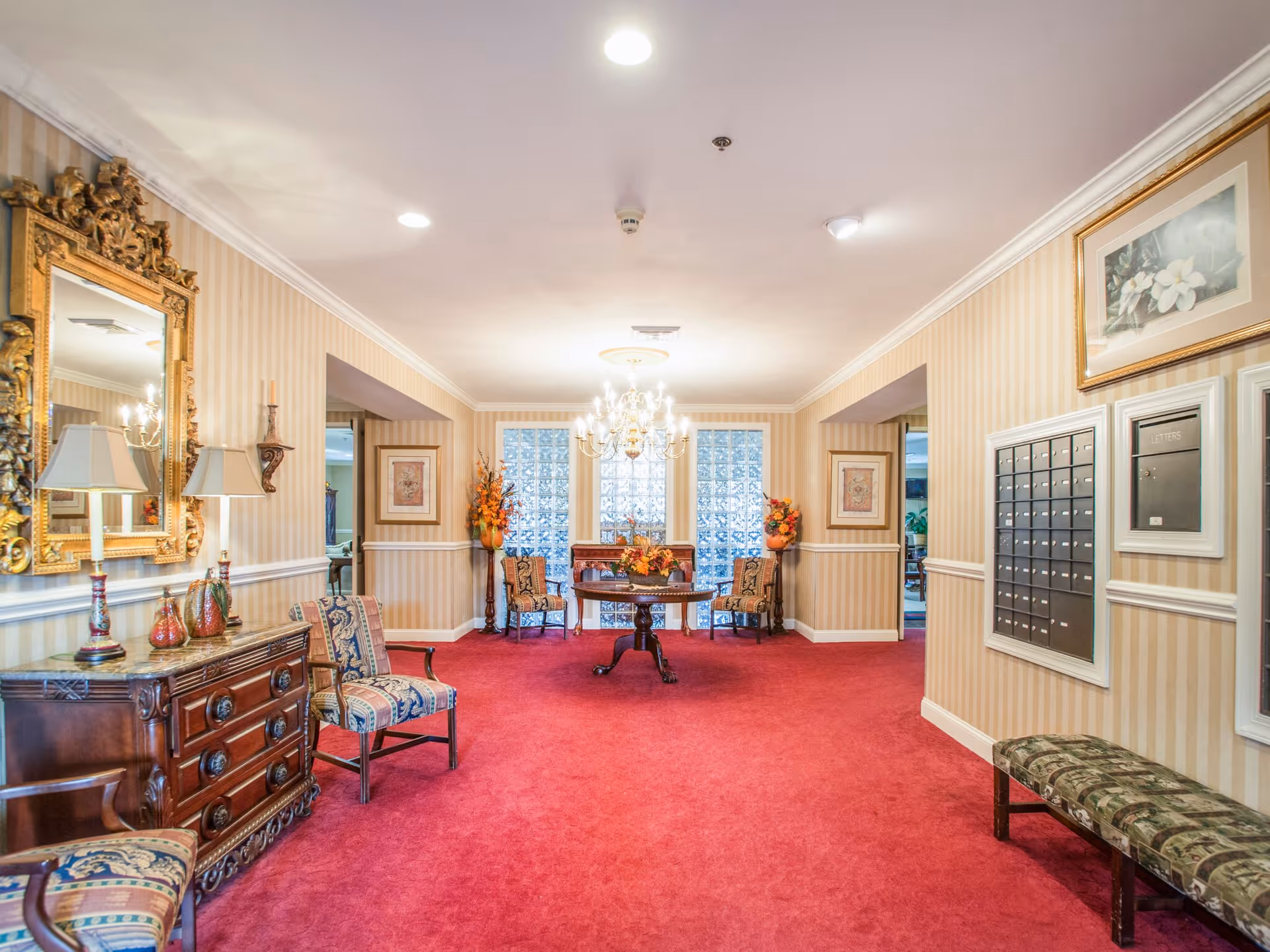 A well-lit interior hallway with red carpet flooring and beige striped wallpaper. The hallway features a wooden dresser with two lamps and decorative items on the left, a large ornate gold-framed mirror above it, and two upholstered chairs with patterned fabric. At the far end, there is a round wooden table with a floral arrangement, flanked by two chairs and two tall vases with orange flowers. On the right wall, there are multiple mailboxes and a framed floral picture above a cushioned bench. A chandelier hangs from the ceiling in the center of the hallway.