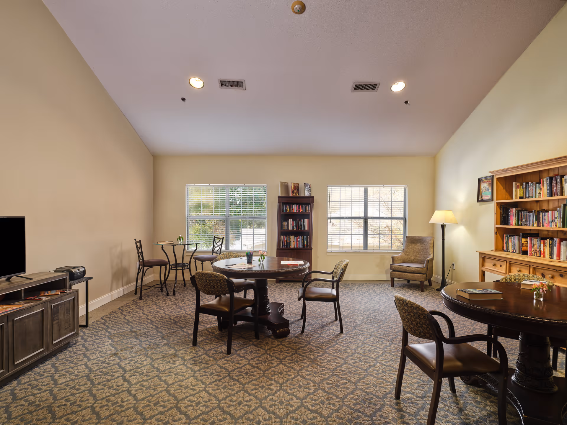 A cozy common area in an assisted living facility featuring patterned carpet, two round wooden tables with chairs, a small round table with two chairs near the window, bookshelves filled with books, a floor lamp, an armchair, and a TV on a wooden stand. The room has beige walls and two large windows letting in natural light.