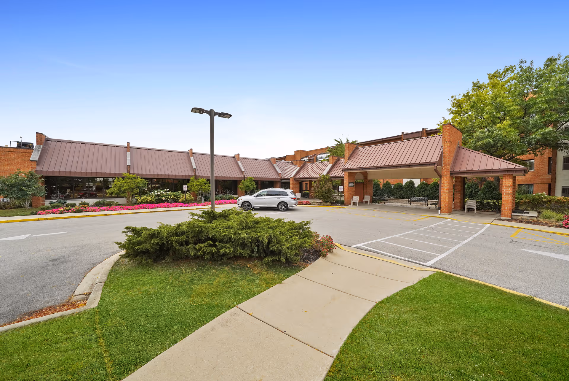 Entrance and covered drop-off driveway of a brick senior living facility with landscaped beds and a parked car.