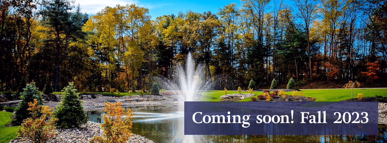 A landscaped pond with a central water fountain, surrounded by trees and fall foliage, overlaid with a banner reading "Coming soon! Fall 2023".