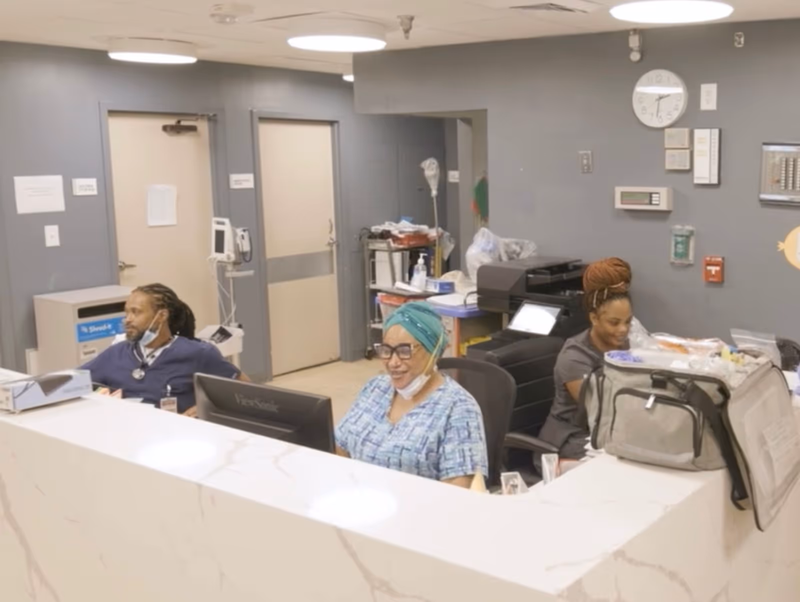 Reception area in a nursing and rehabilitation center with three staff members seated behind a white marble counter. The background shows two closed doors, medical equipment, and office supplies. The walls are gray with a clock and various control panels mounted.