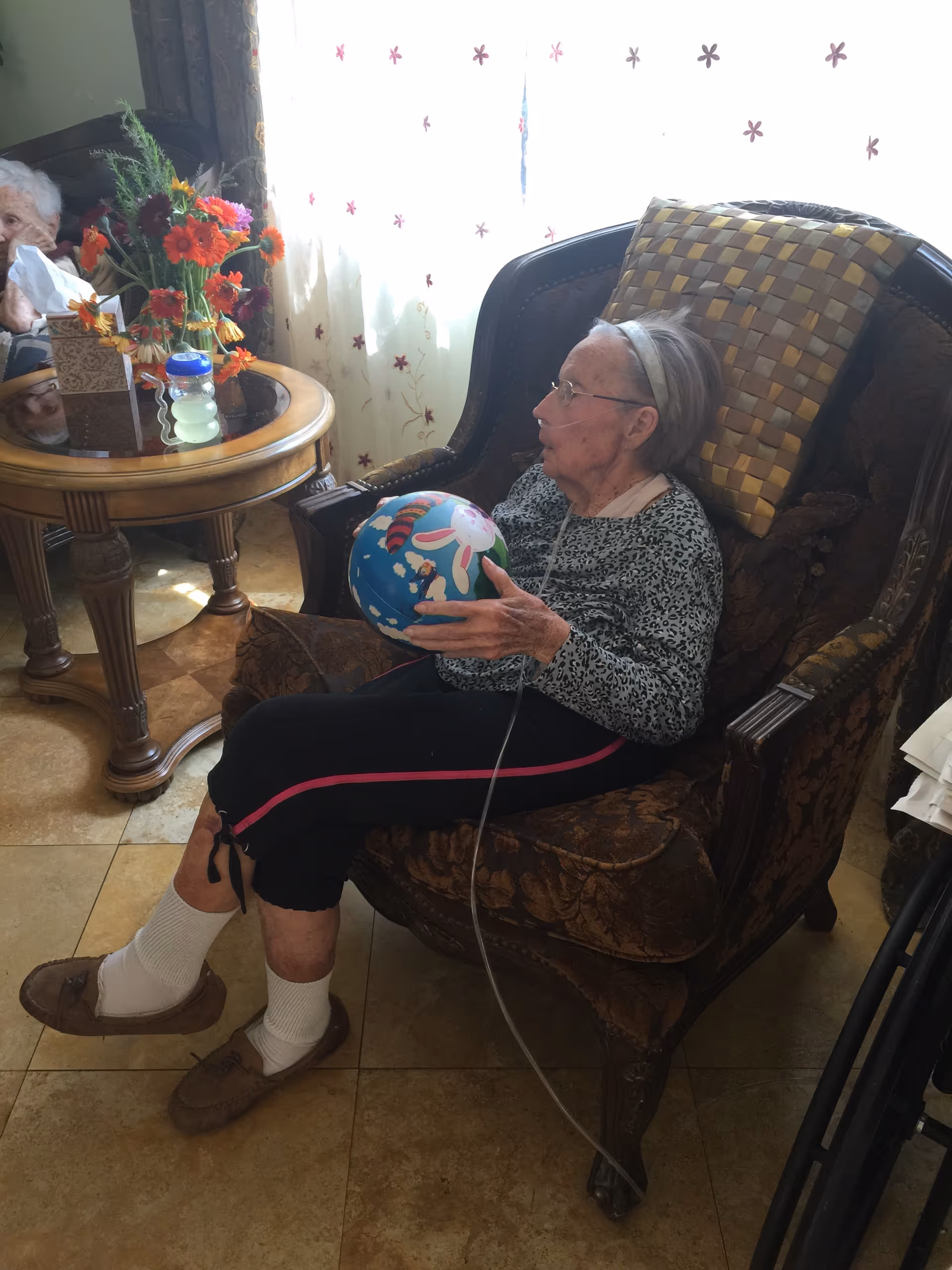 An elderly woman sits in an upholstered armchair holding a colorful ball in a sunlit sitting area with a side table and flowers.