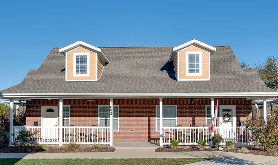 Front exterior of a brick single-story building with a covered porch, white railing, and two dormer windows under a clear blue sky.
