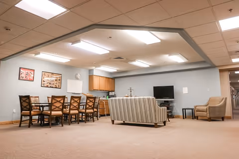 A spacious common area in a senior living facility featuring a dining table with eight chairs, a striped sofa facing a flat-screen TV on a stand, a single armchair, and a kitchenette with wooden cabinets. The walls are light blue, and the ceiling has recessed lighting panels.