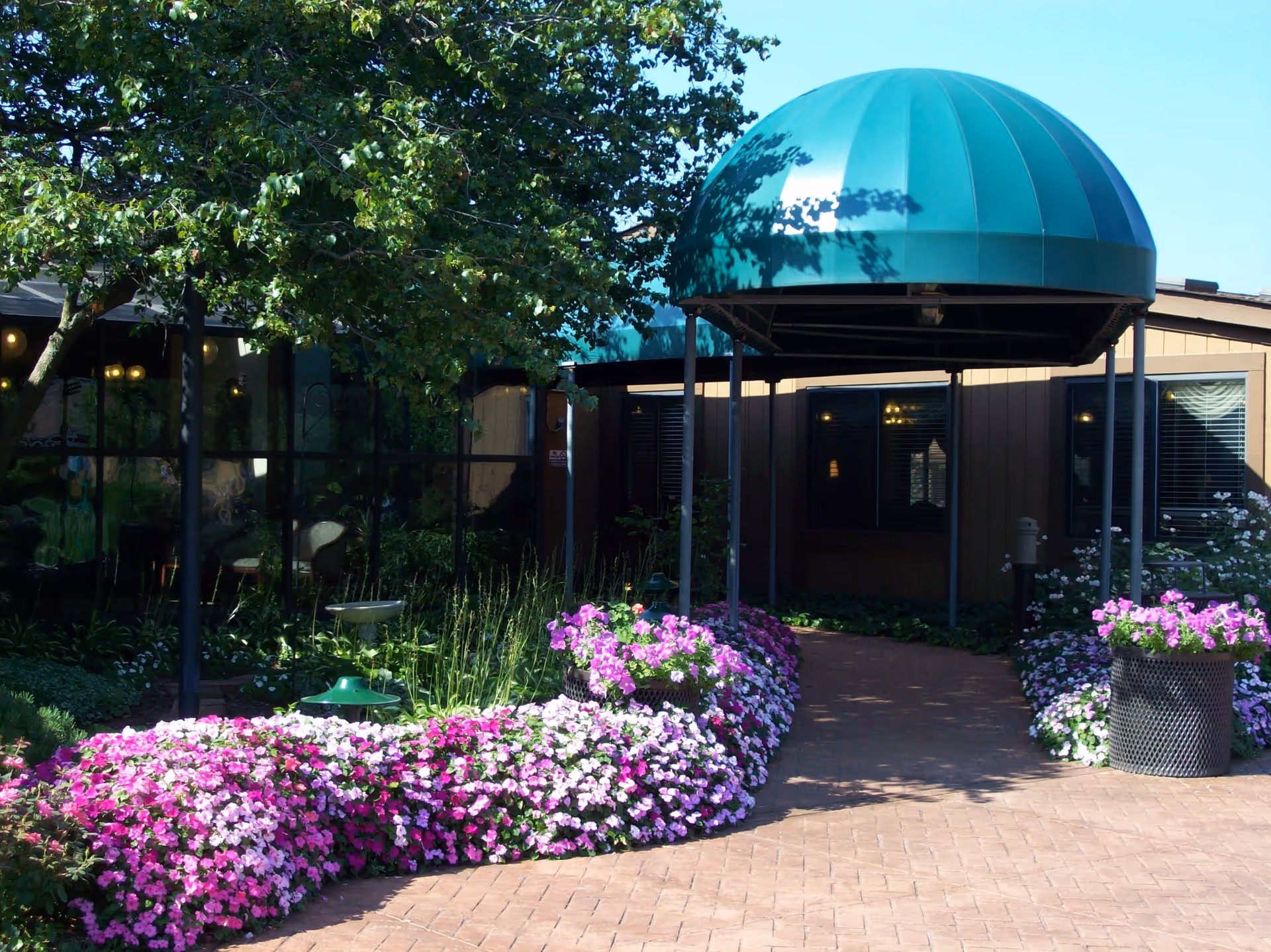 Entrance to a building with a green dome-shaped canopy over a brick pathway, surrounded by vibrant flower beds with pink, purple, and white flowers, and a tree providing shade on the left side.