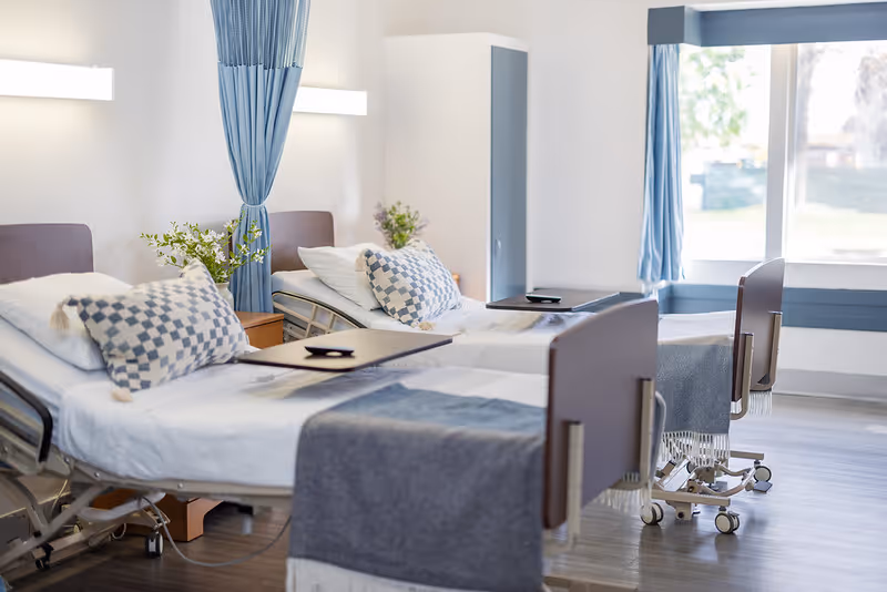 A bright and clean nursing facility room with two hospital beds, each with a checkered pillow and a blue-gray blanket. There are bedside tables with flowers, a blue curtain between the beds, and a large window with blue curtains letting in natural light.
