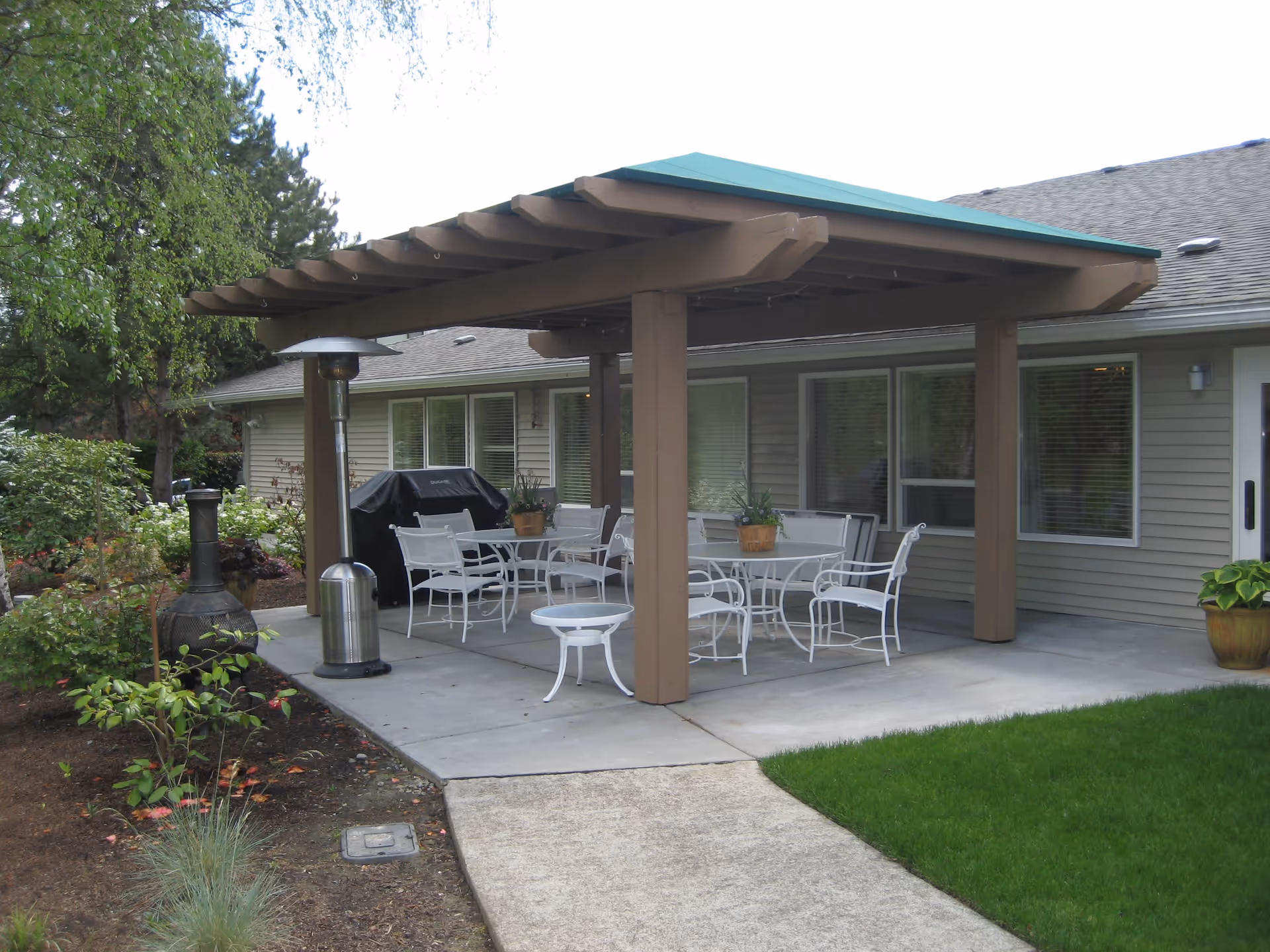 Outdoor patio area at Aegis Living Redmond with a wooden pergola covering white metal tables and chairs. There are potted plants on the tables, a grill covered with a black cover, a patio heater, and a chiminea. The patio is surrounded by green grass and garden plants, adjacent to the building with beige siding and multiple windows.