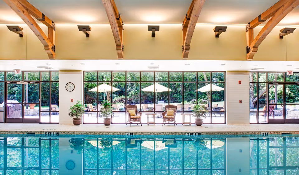 Indoor swimming pool area with clear blue water, wooden ceiling beams, large windows showing outdoor patio with umbrellas and chairs, potted plants, and a clock on the wall.