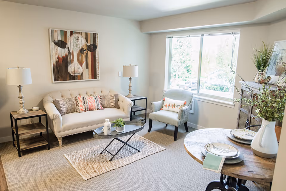 A bright and cozy living room with a beige tufted sofa adorned with patterned pillows, a light gray armchair with a decorative pillow, two side tables each with a lamp, a small oval coffee table with a plant and decorative items, a round wooden dining table set with plates and napkins, a large window letting in natural light, and a colorful cow painting on the wall.