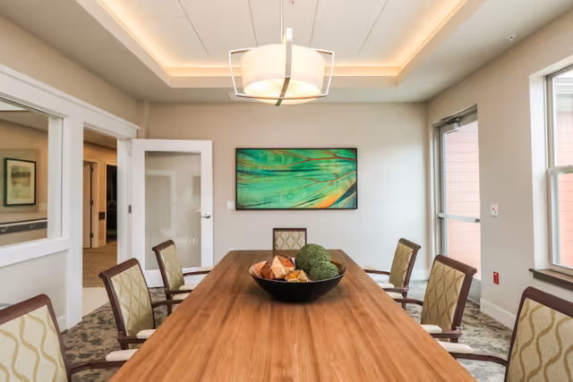 A well-lit dining room with a long wooden table surrounded by eight cushioned chairs. A modern light fixture hangs from the ceiling above the table. On the table, there is a decorative bowl with ornamental spheres. The room has beige walls, a large window on the right, a glass door, and a colorful abstract painting on the far wall.
