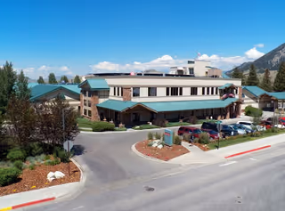 Exterior view of St. John's Living Center, a multi-story building with green roofs, surrounded by trees and parked cars under a clear blue sky with mountains in the background.