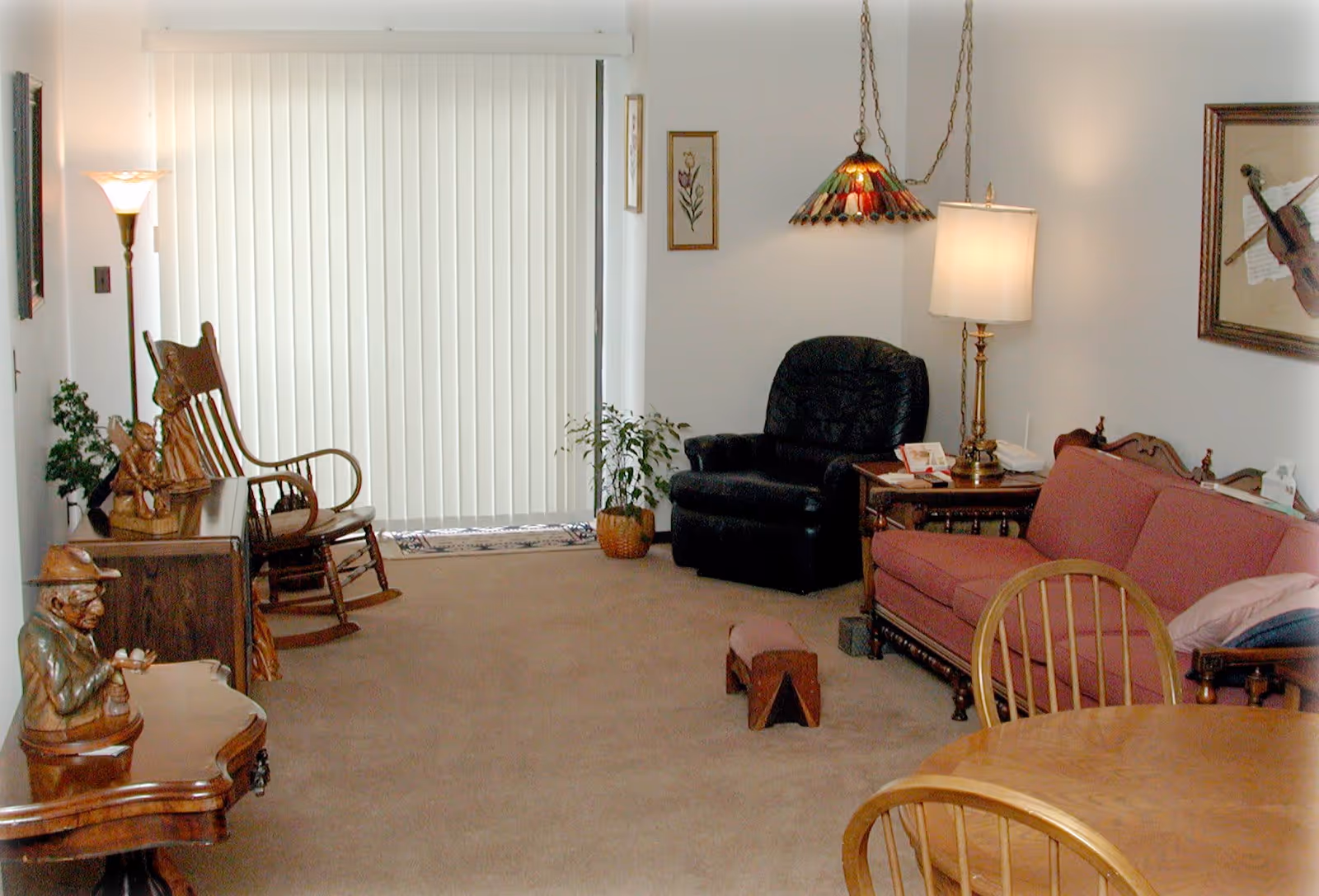 A cozy living room with a red sofa, black recliner chair, wooden rocking chair, wooden side tables, a floor lamp, a hanging stained glass lamp, framed artwork on the walls, and vertical blinds covering a sliding glass door.