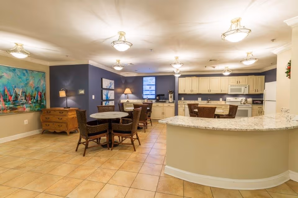 A spacious kitchen and dining area in a senior living facility with beige tiled floors, cream-colored cabinets, a granite countertop island, and several round tables with wicker chairs. The walls are painted in a combination of beige and dark blue, decorated with colorful artwork and a lamp on a wooden dresser.
