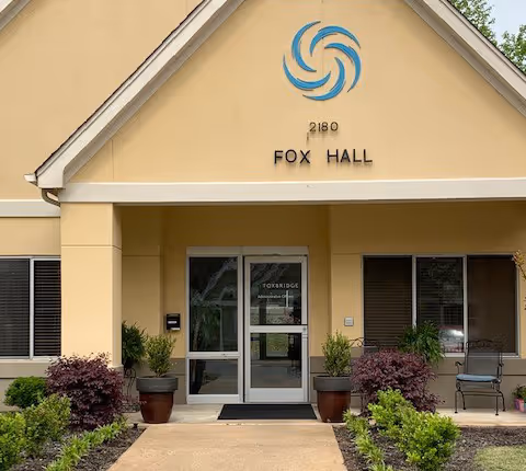Entrance to the Fox Hall building at Foxbridge with a beige facade, glass double doors, potted plants, and signage reading 'FOX HALL' and '2180'.