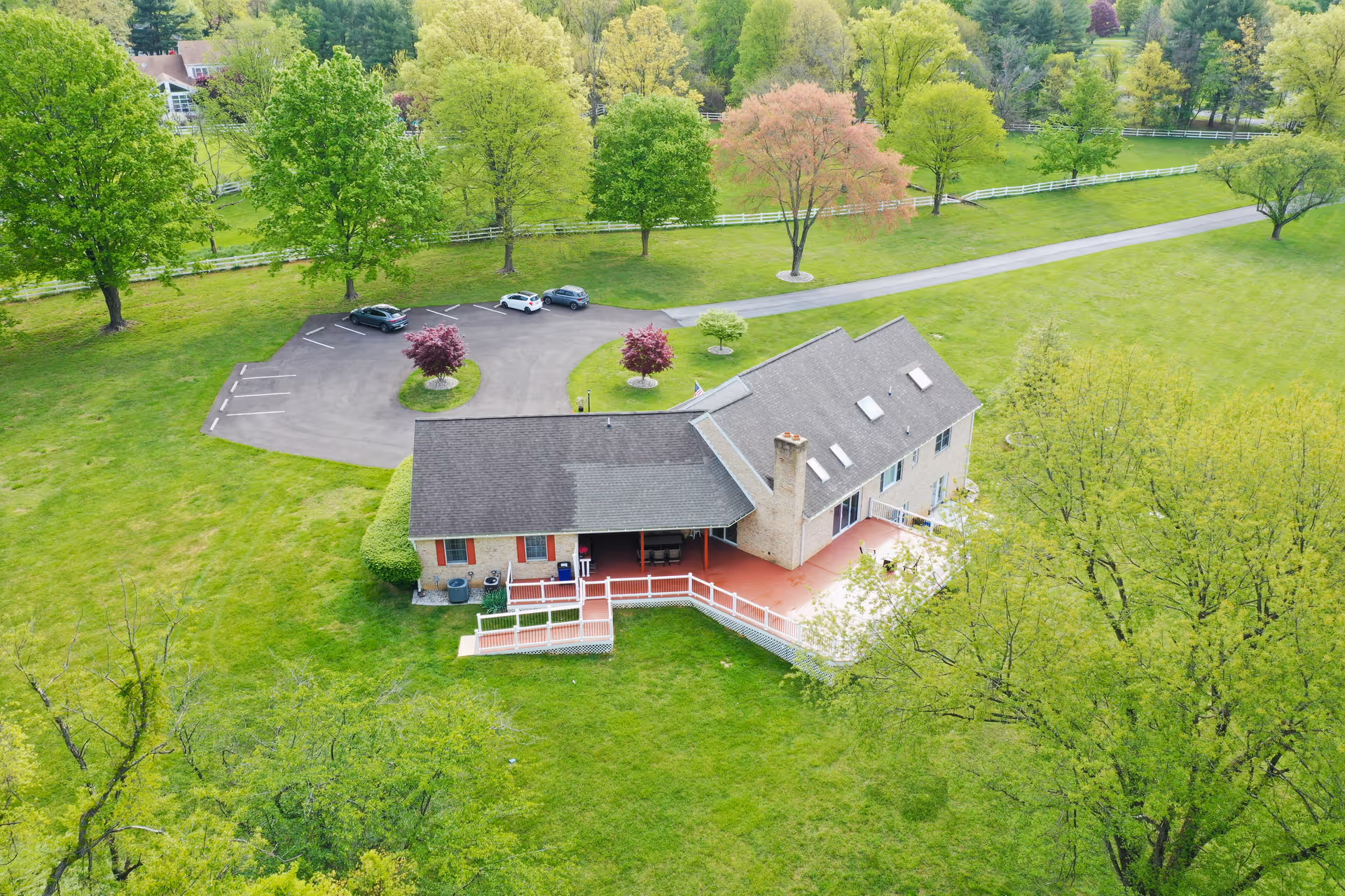 Aerial view of a large residential building surrounded by green grass and trees. The building has a gray roof, a chimney, and a red patio area with white railings. There is a small parking lot with a few cars and landscaped islands with trees. The area is spacious and lush with greenery.
