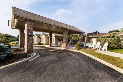 Covered porte-cochère entrance of The Pines Senior Living with brick columns, white outdoor chairs, and landscaped plantings.