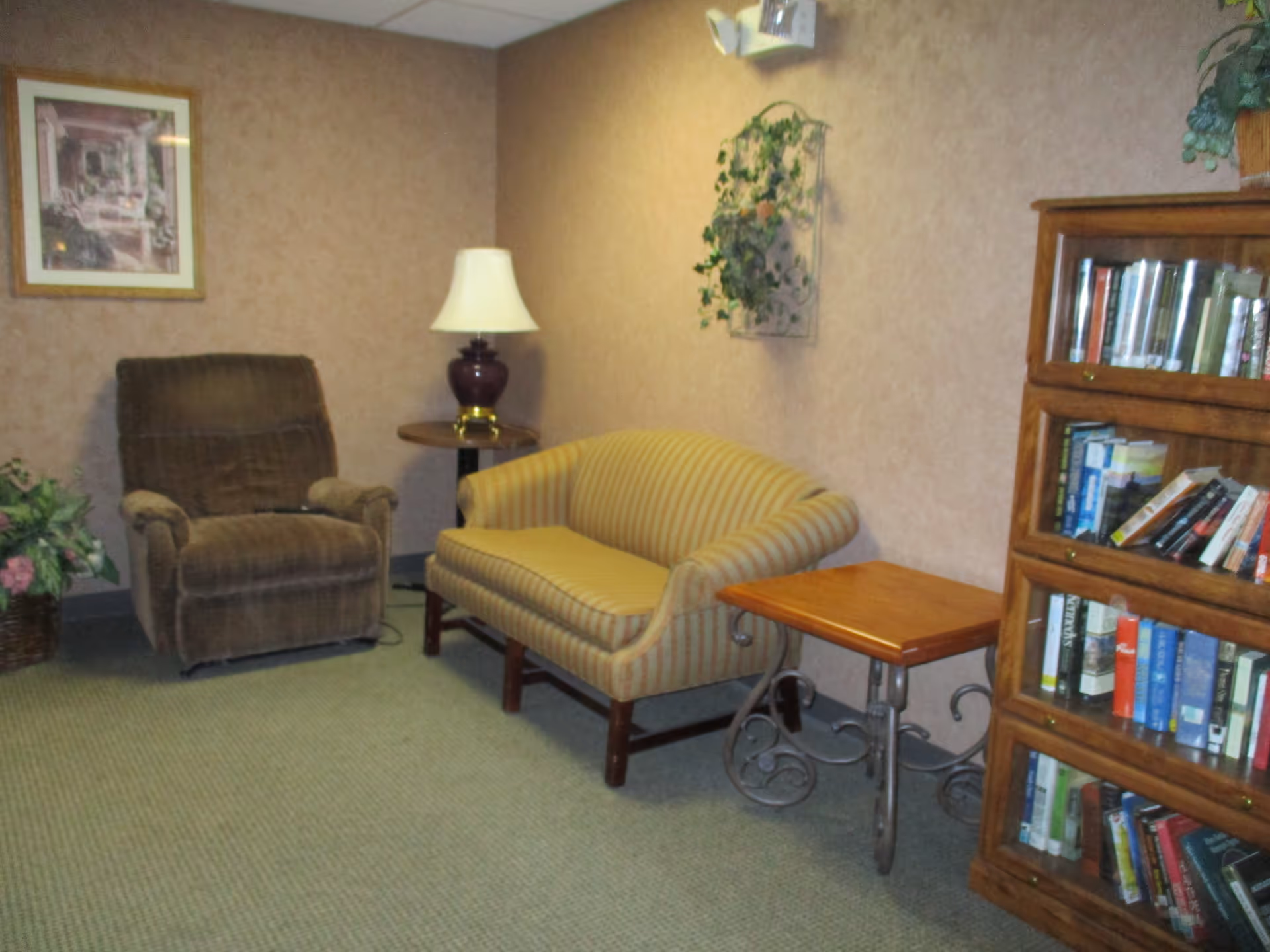 A cozy sitting area with a brown recliner, striped loveseat, side tables, lamp, wall decor, and a bookshelf of books.