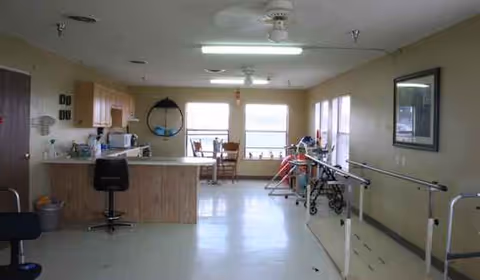 Interior rehab room with a reception desk, therapy bars, mobility aids, and chairs.