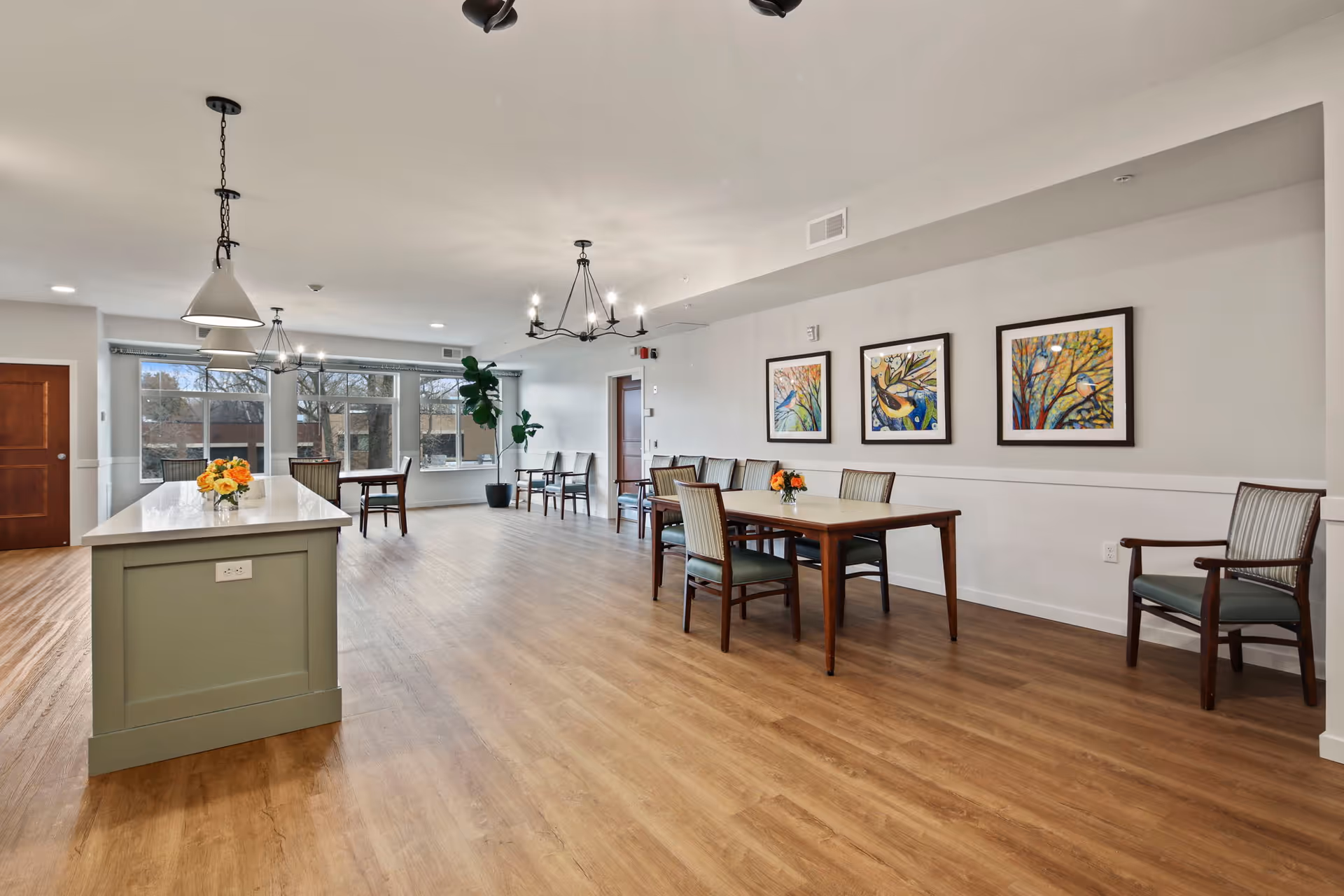 A spacious, well-lit common area with wooden flooring and light-colored walls. The room features a large kitchen island with pendant lights above it, a wooden dining table with chairs, and additional seating along the walls. Three colorful framed artworks hang on the wall, and large windows allow natural light to fill the space. A potted plant is placed near the windows.