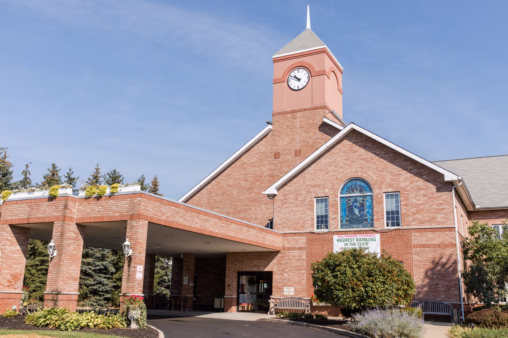 Exterior view of Hillebrand Nursing & Rehabilitation Center, a brick building with a clock tower, a covered entrance, and landscaping including bushes and flowers under a clear blue sky.