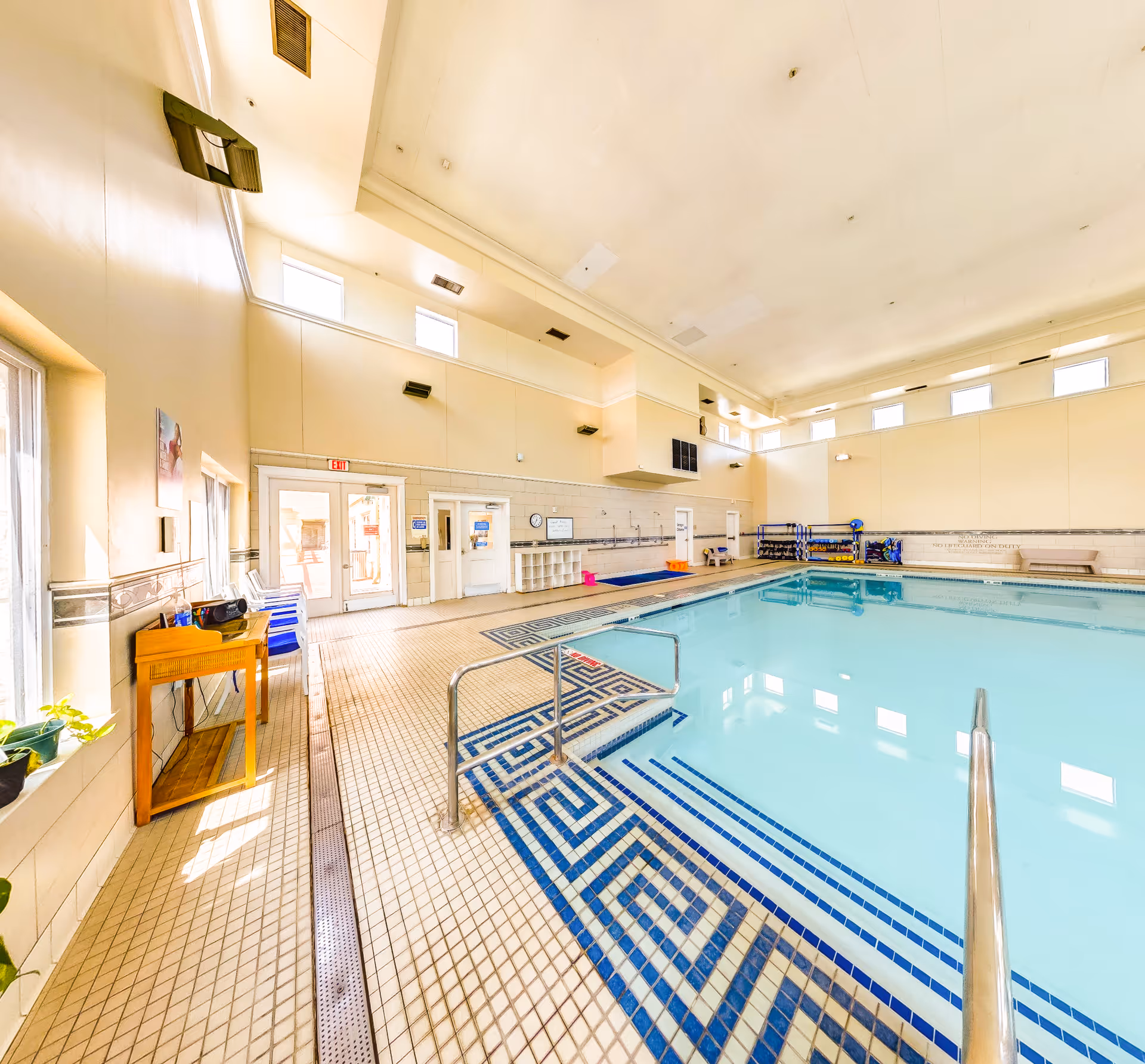 Indoor swimming pool area with tiled floor and walls, metal handrails leading into the pool, several windows near the ceiling allowing natural light, a wooden table with chairs on the left side, and pool equipment stored along the far wall.