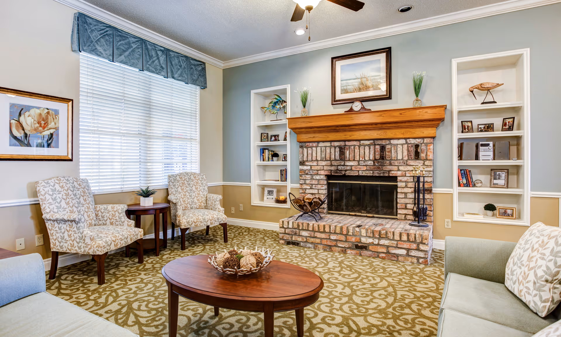 A cozy living room with a brick fireplace centered on the wall, flanked by built-in white shelves holding decorative items and framed photos. Two patterned armchairs are placed near a window with white blinds and a blue valance. A wooden coffee table with decorative balls sits on a patterned carpet, and a light green sofa with a patterned pillow is partially visible.