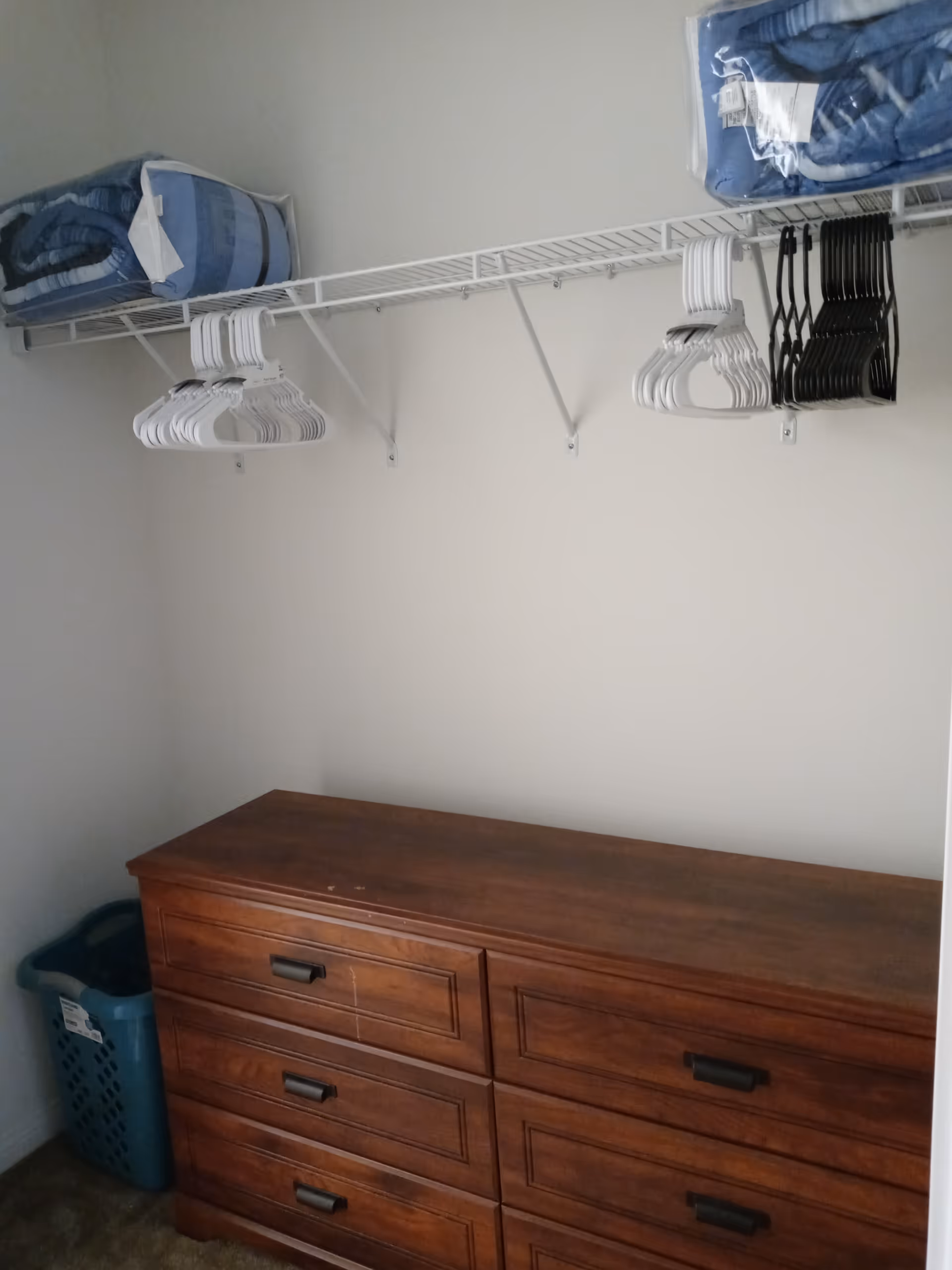 A closet space with a wooden dresser featuring six drawers, a blue laundry basket in the corner, and a white wire shelf above holding several white and black hangers and two folded blue blankets in plastic packaging.