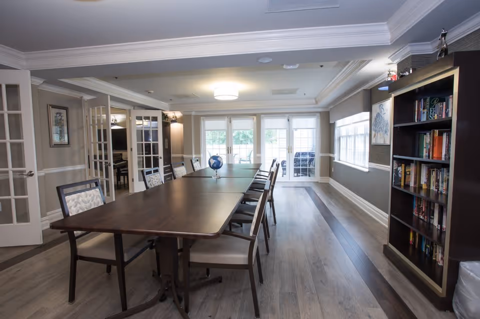 A well-lit room with a long dark wooden table surrounded by chairs, a small globe on the table, a bookshelf filled with books on the right, and large windows and glass doors letting in natural light at the far end.