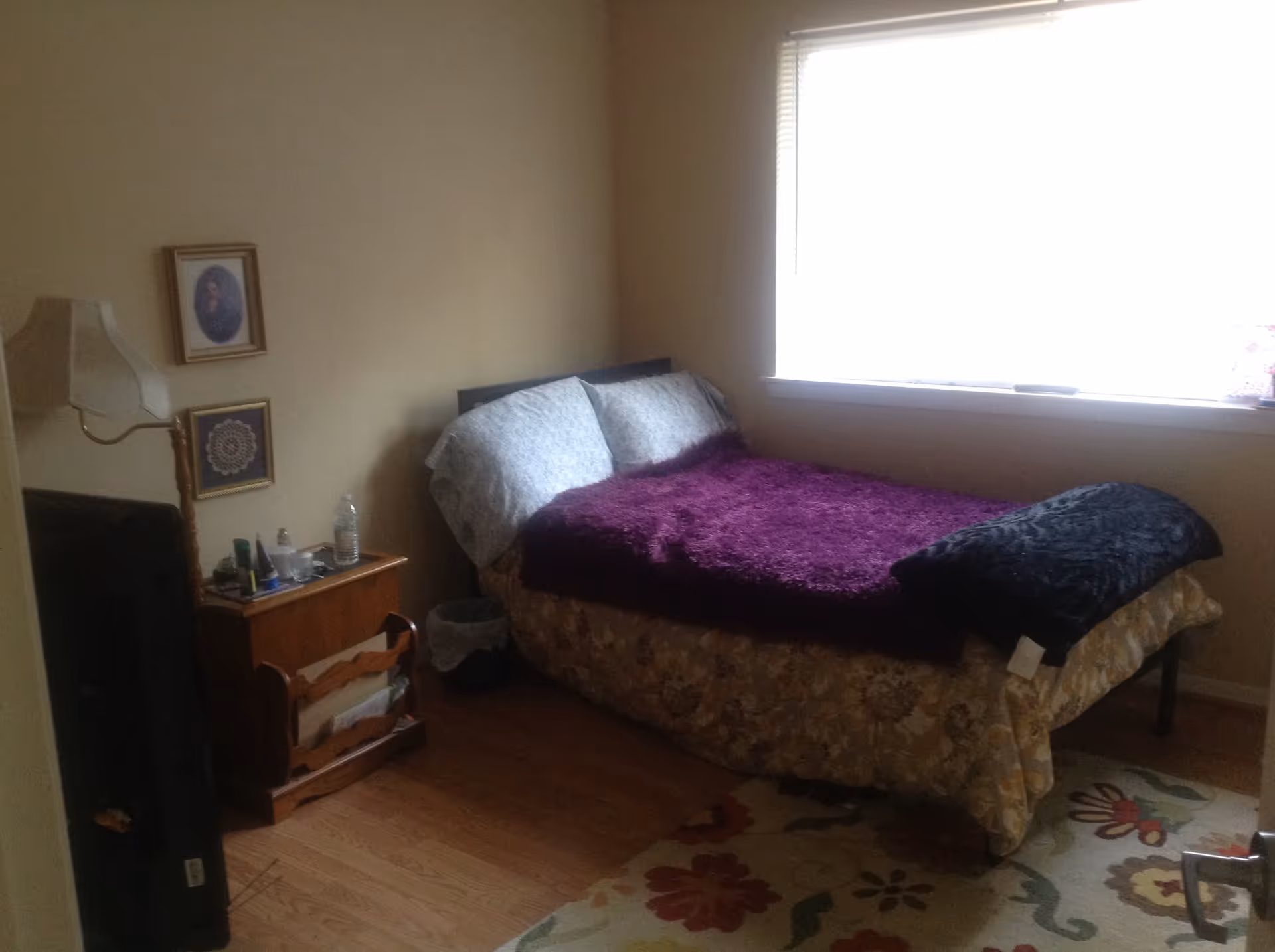 A small bedroom with a bed covered in a floral bedspread and a purple blanket. There are two pillows on the bed. Next to the bed is a wooden nightstand with various items including a water bottle and a lamp. Two framed pictures hang on the wall above the nightstand. A window with blinds is on the right side, letting in natural light. The floor is wooden with a floral area rug partially visible.