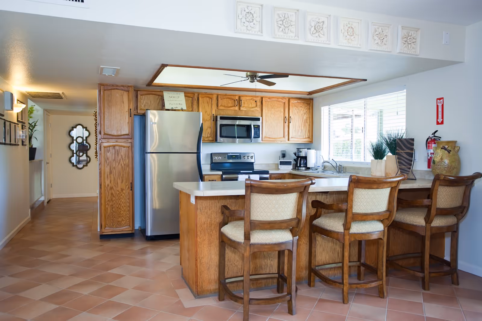 A bright kitchen area with wooden cabinets, a stainless steel refrigerator, microwave, and stove. There is a kitchen island with a beige countertop and three wooden chairs with cushioned seats. A window with blinds lets in natural light, and decorative items including plants and a fire extinguisher are placed on the countertop. The floor is tiled with a warm-toned pattern.