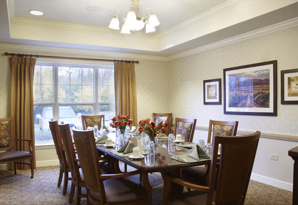 Formal dining room with a large table set for a meal, floral centerpieces, wooden chairs, and a window with curtains.