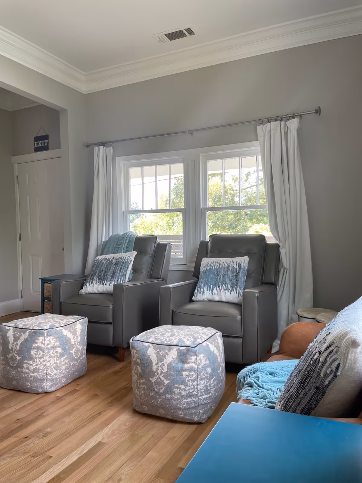 A cozy living room with two gray armchairs each adorned with a decorative pillow, two patterned ottomans in front of the chairs, a brown leather chair with a blue throw blanket and pillow, light wood flooring, and two windows with white curtains letting in natural light.