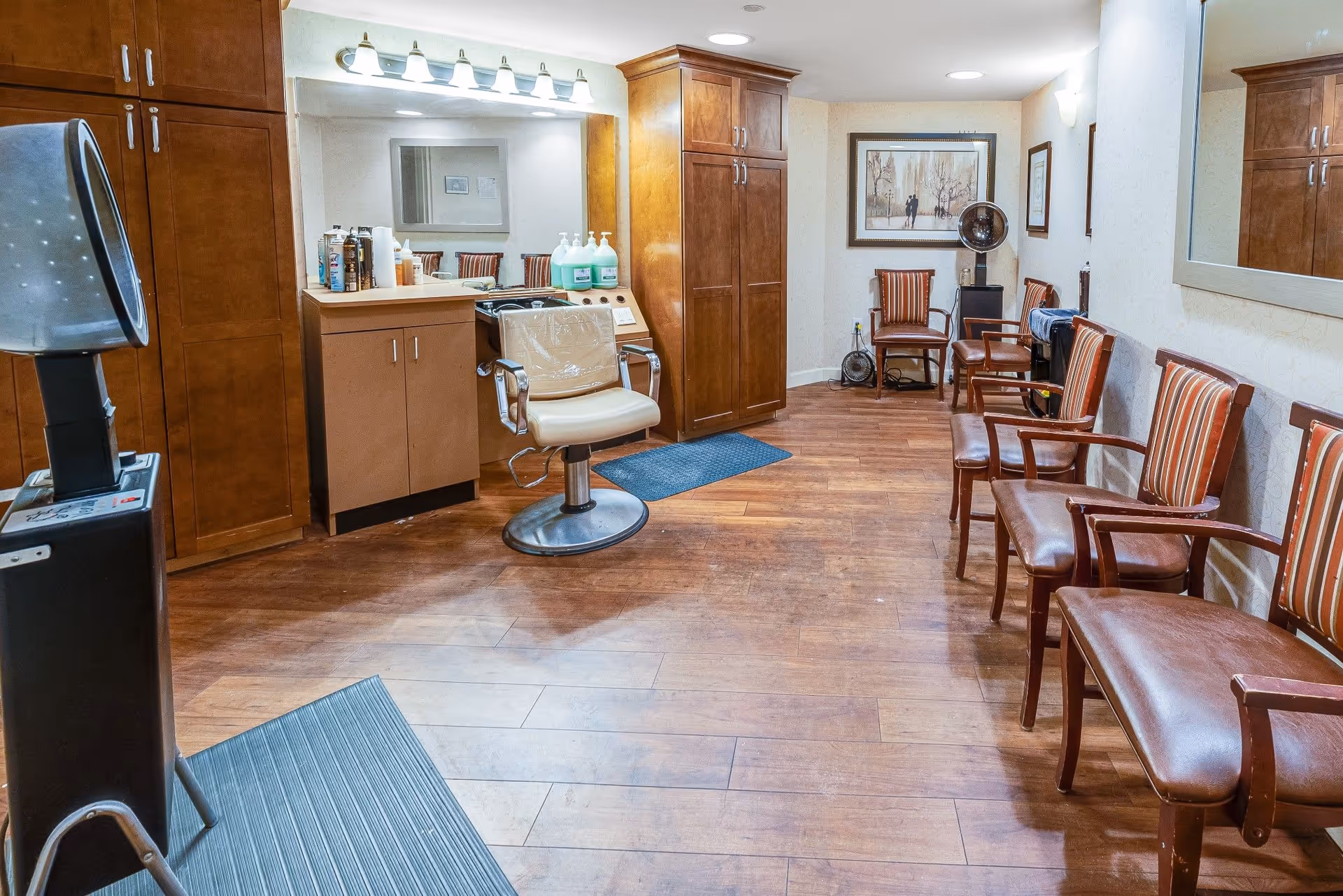 Interior view of a salon area in a senior living facility with wooden flooring, a styling chair in front of a counter with hair care products, a hair dryer station on the left, and several striped upholstered chairs lined up against the right wall. The room has wooden cabinets, framed artwork on the walls, and bright overhead lighting.