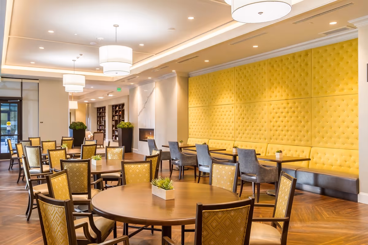 Bright communal dining room with round wooden tables, yellow tufted banquette seating, and pendant lighting.