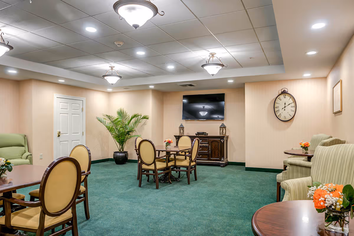 A cozy common area with green carpet and beige walls featuring multiple seating arrangements including round wooden tables with cushioned chairs and armchairs. A flat-screen TV is mounted on the wall above a wooden cabinet, and a large clock is hung on the adjacent wall. The room is softly lit by ceiling lights and decorative hanging fixtures, with a potted plant adding greenery in the corner.