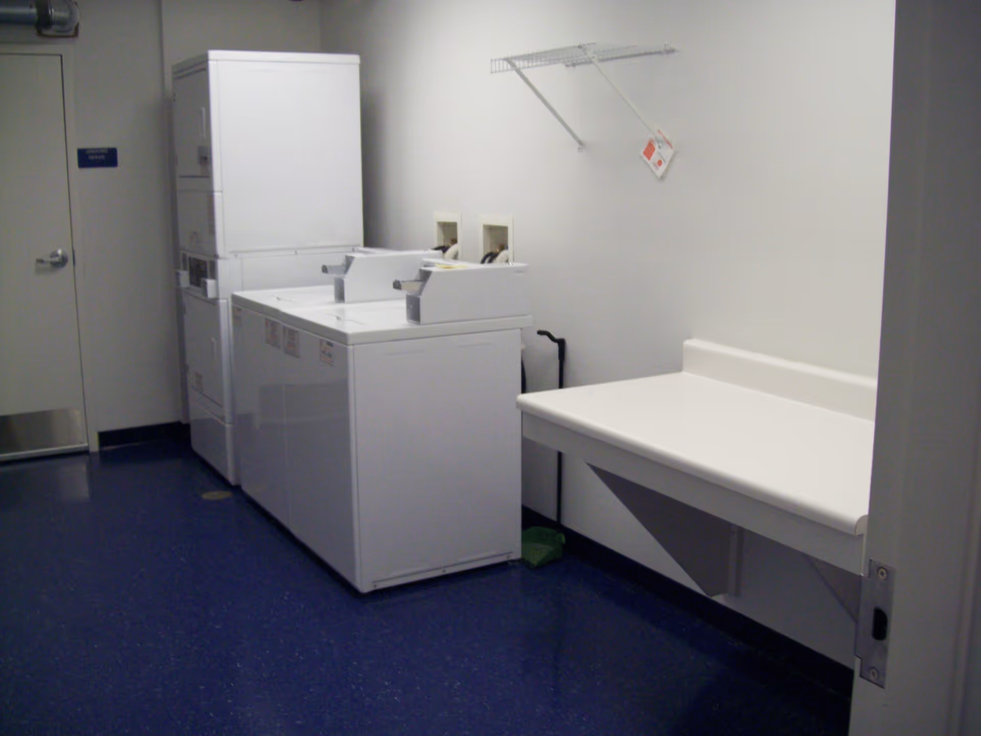 Laundry room with two white top-loading washing machines, a stacked washer and dryer unit, a folding table attached to the wall, and a wire shelf above the table. The floor is blue and the walls are white.