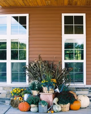 Exterior view of a building with brown siding and large white-framed windows. In front of the building, there is a decorative autumn display featuring pumpkins, gourds, potted flowers, and dried corn stalks arranged on a stone ledge.