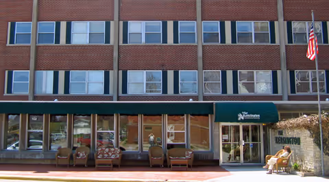 Front facade of a multi-story brick senior living building with a green awning reading The Kensington, entrance doors, several chairs on the sidewalk, and an American flag.