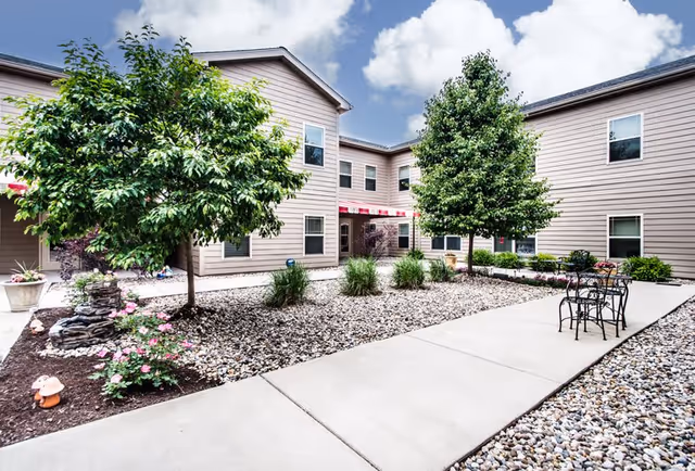 Outdoor courtyard area of a senior living facility with a concrete walkway, small trees, shrubs, and decorative rocks. The building is two stories with beige siding and several windows. There is a small black metal table with two chairs on the right side of the walkway. The sky is partly cloudy.