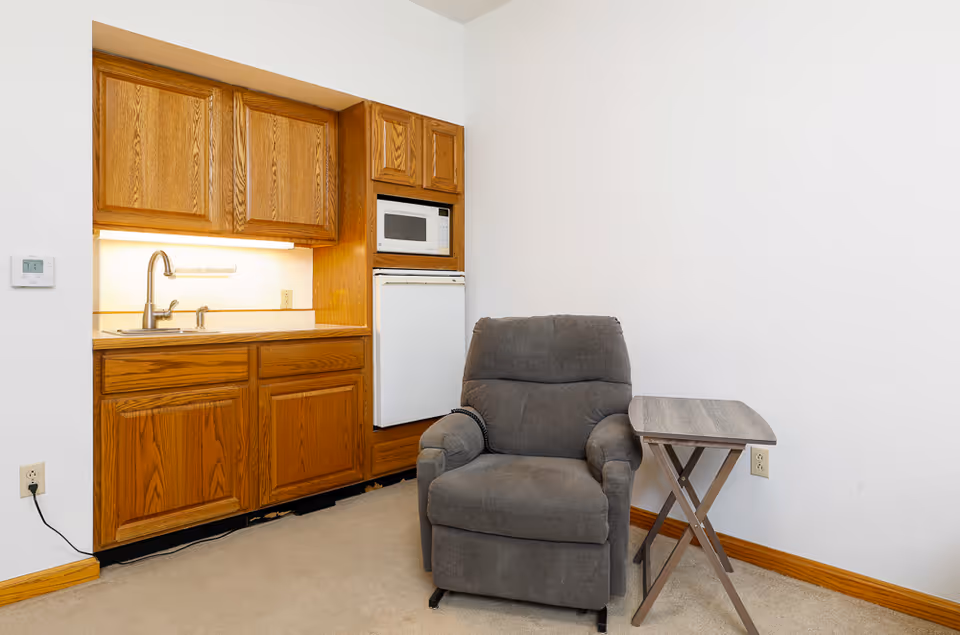 Small living area with a gray recliner and side table beside a kitchenette with oak cabinets, sink, microwave, and compact refrigerator.