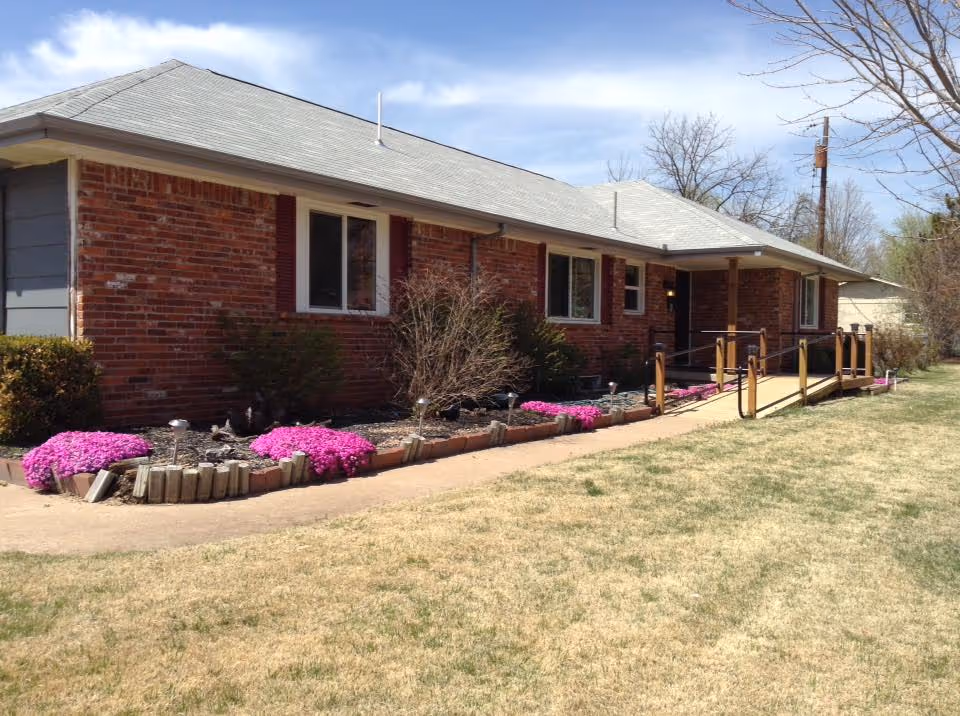 Single-story brick building with a gray roof, several windows, and a wooden ramp leading to the entrance. The front yard has a pathway bordered by flower beds with pink flowers and small bushes. The sky is partly cloudy and there are leafless trees in the background.