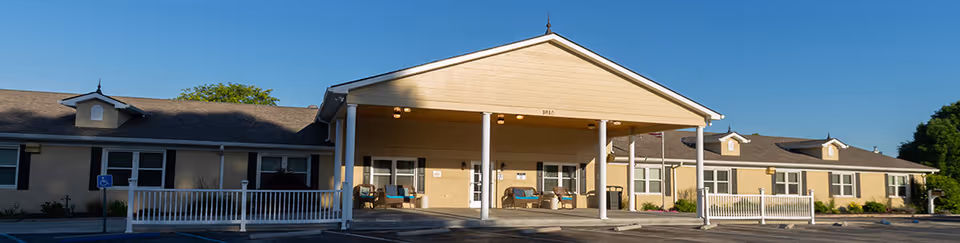 Exterior view of Brownsburg Health Care Center, a single-story building with a covered entrance supported by white columns. The building has beige walls, multiple windows, and a parking area in front with a handicap parking sign visible. The sky is clear and blue.
