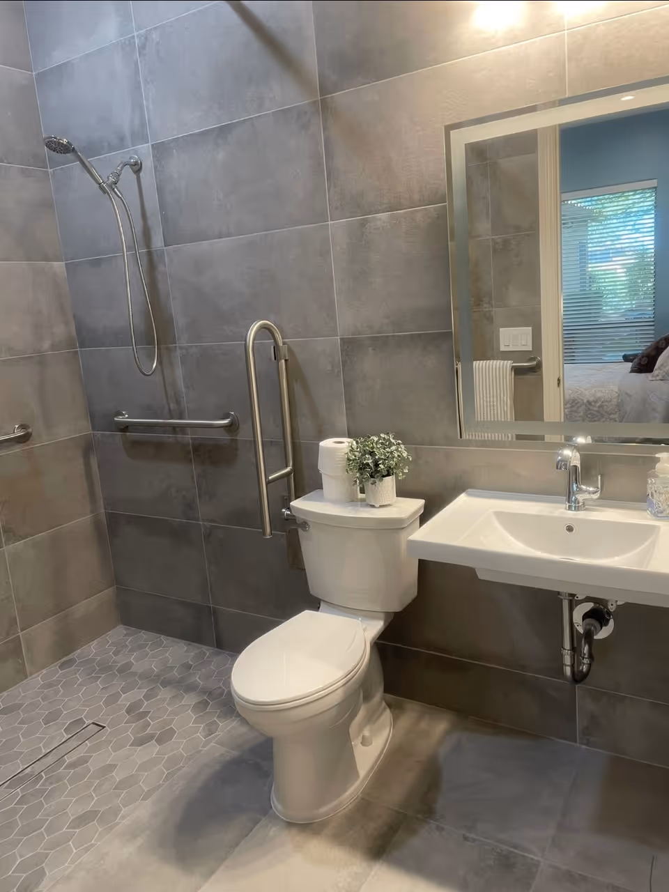 A modern bathroom with gray tiled walls and floor. It features a white toilet with a roll of toilet paper and a small potted plant on top. There is a wall-mounted white sink with a chrome faucet and a soap dispenser. A large mirror is mounted above the sink, reflecting part of a bedroom with a window and bed. The shower area has a handheld showerhead and grab bars for accessibility.