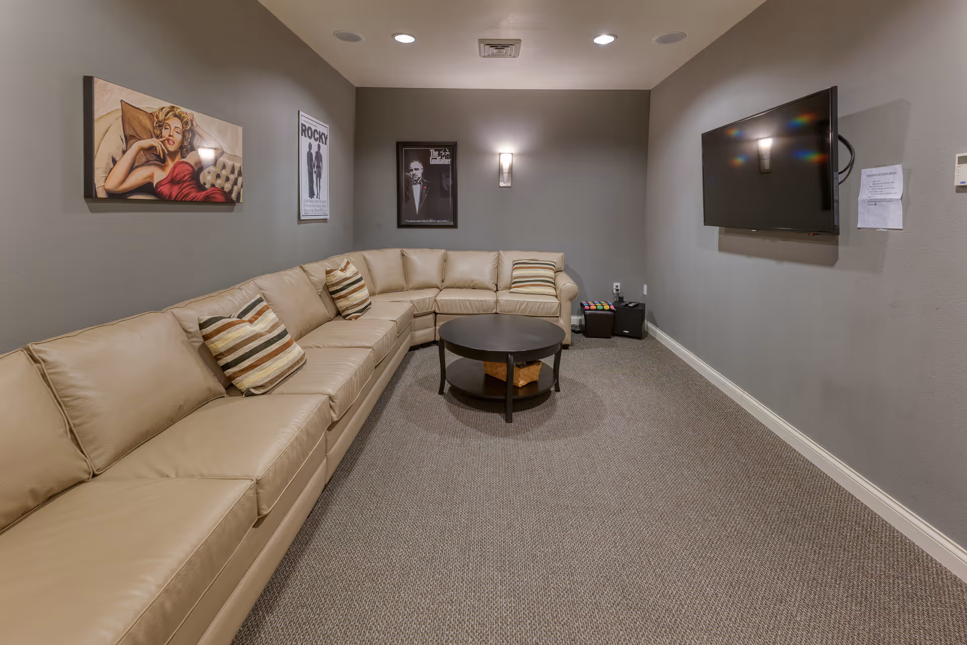 A cozy lounge with a beige sectional sofa, round coffee table, wall-mounted TV, and framed artwork on gray walls.