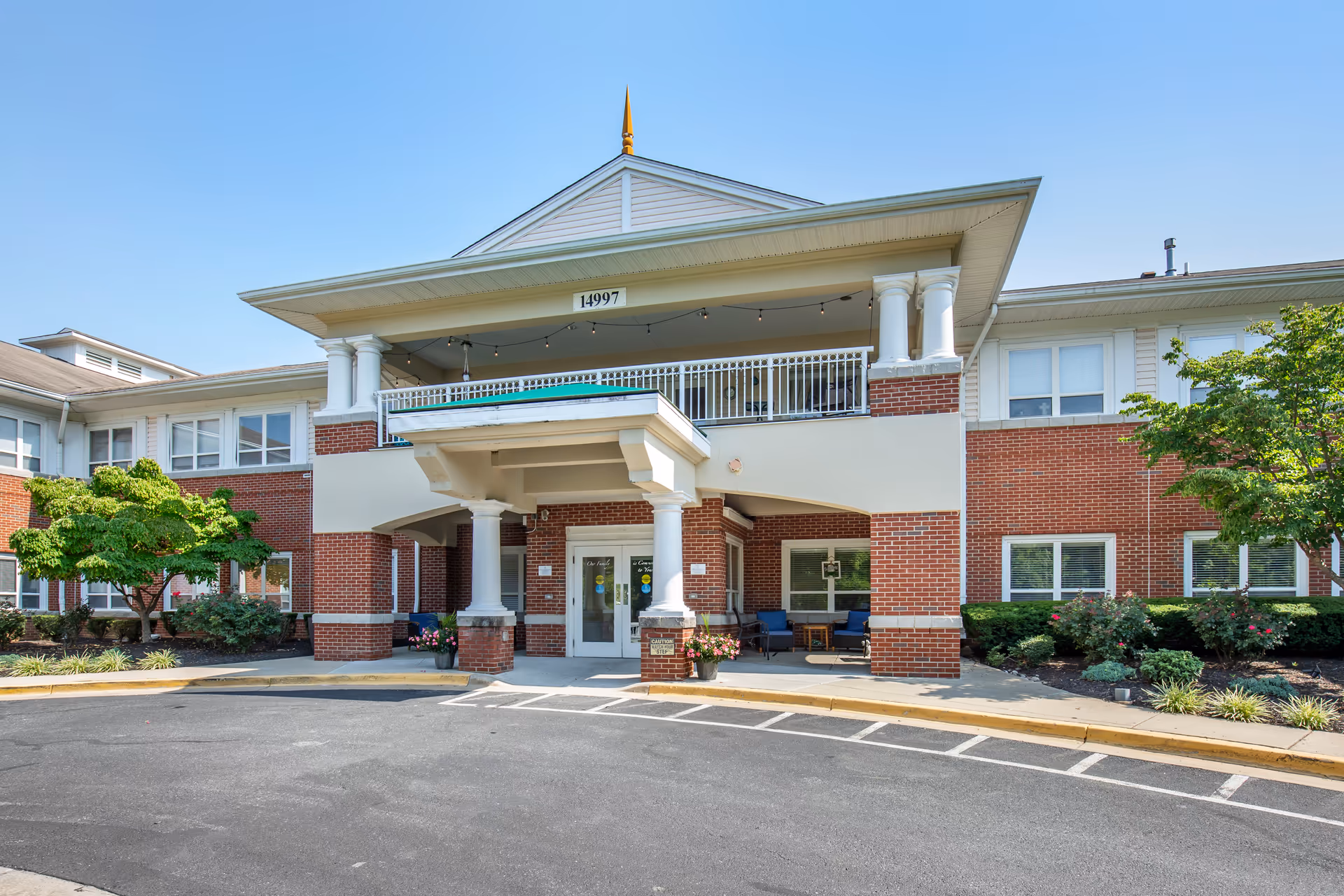 Front exterior view of Brookdale Woodward Estates, a two-story brick building with white columns and a covered entrance. There are trees and shrubs on either side of the entrance, and a clear blue sky overhead.