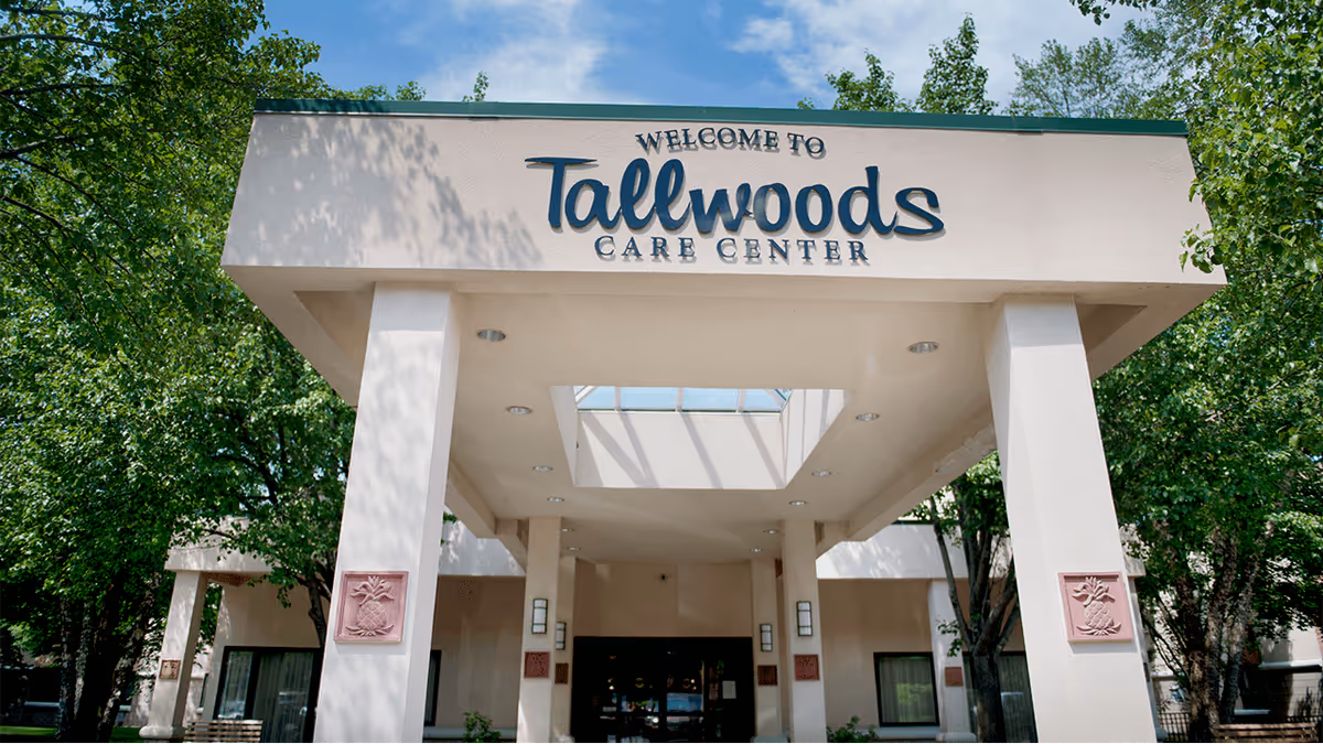 Entrance of Tallwoods Care Center with a covered walkway supported by columns, surrounded by green trees under a blue sky.