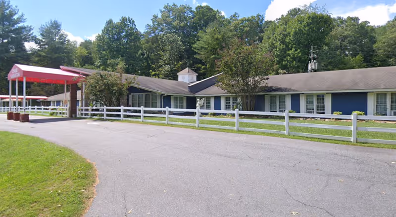 Single-story assisted living building with a covered red entrance canopy, white picket fence, and trees in the background.