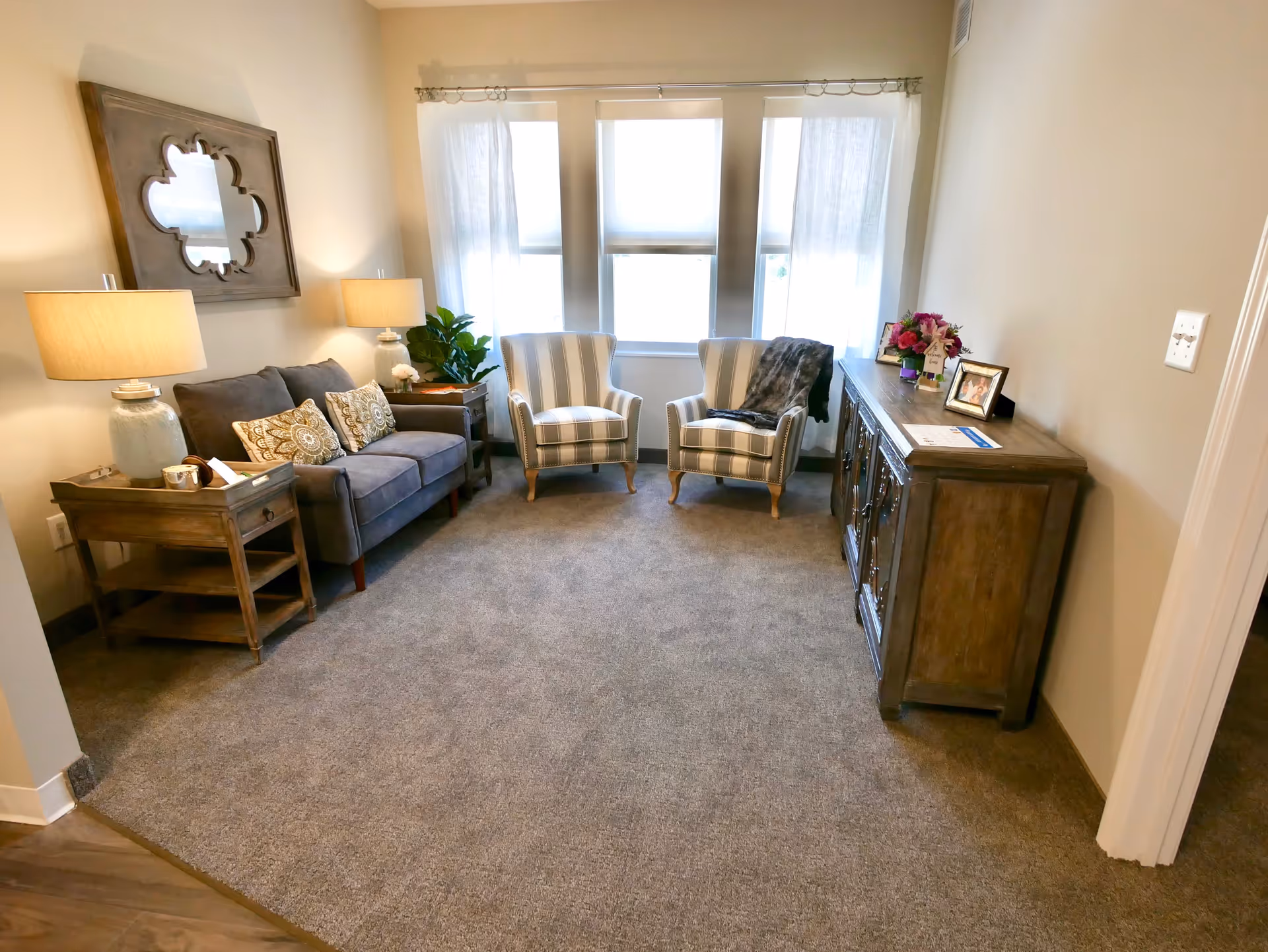 A cozy living room with a gray sofa adorned with patterned cushions, two striped armchairs, a wooden side table with a lamp, a wooden cabinet with framed photos and flowers on top, and a large window with sheer curtains letting in natural light.