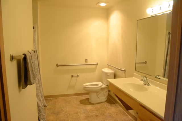 Accessible bathroom with a toilet, vanity sink, mirror, grab bars, towel rack, and tiled floor.