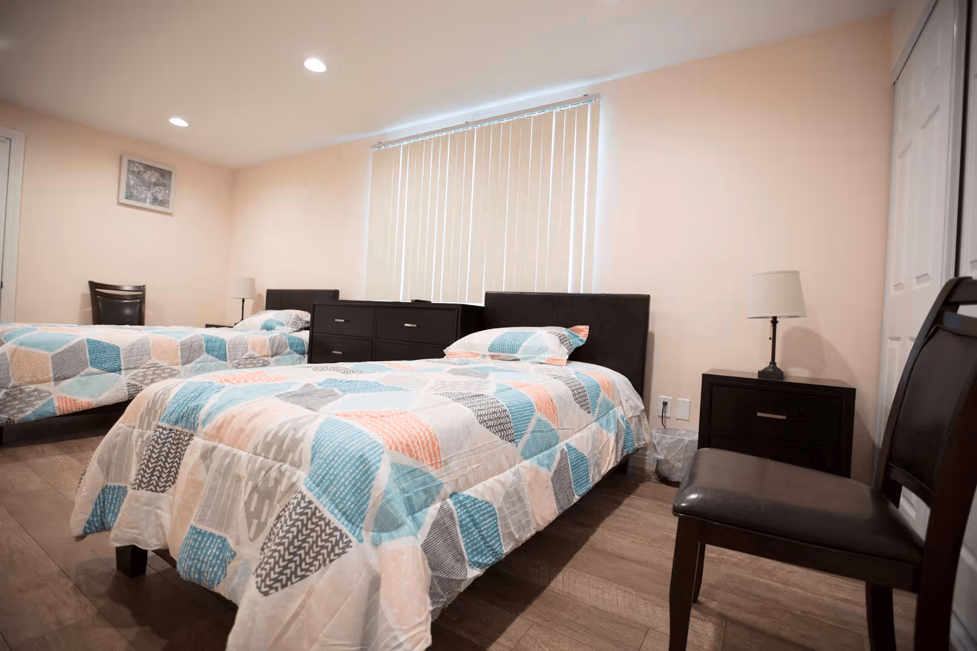 A bedroom with two single beds featuring colorful geometric patterned bedspreads. Between the beds is a dark wooden dresser, and each bed has a matching dark wooden nightstand with a lamp. There is a chair in the foreground and vertical blinds covering the window on the far wall. The room has light-colored walls and wooden flooring.